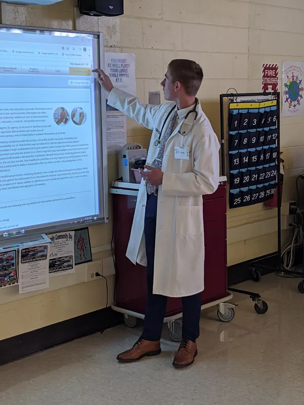 Male student in white lab coat stands in front of large touchscreen board